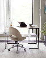 Modern minimalist home office setup with a sleek, white rectangular desk featuring a thin tabletop on black metal legs, paired with a stylish, ergonomic office chair upholstered in a light beige fabric with a gently curved backrest and a chrome 5-star base with casters. Beside the desk is a small, square side table with a black metal frame and a clear glass top, holding a stack of books. The room is complemented by a soft grey area rug and ambient natural light filtering through sheer curtains.