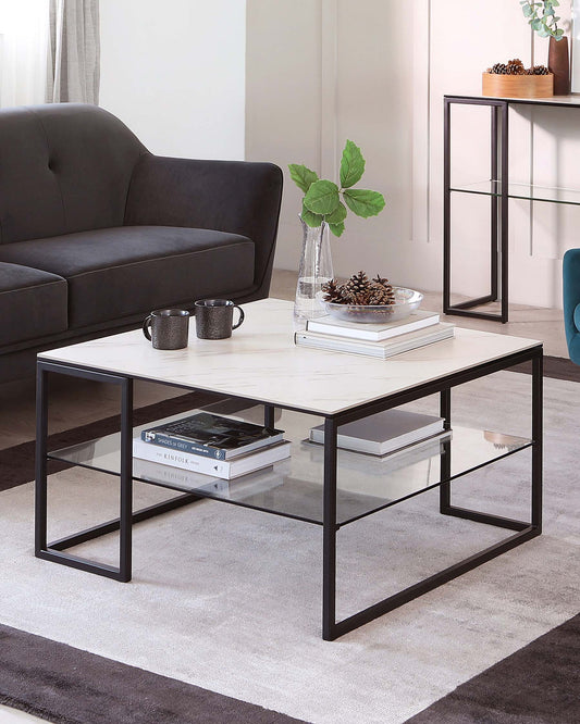 Contemporary living room furniture arrangement featuring a minimalist rectangular coffee table with a white marble-like top and a sleek black metal frame. In the background, a dark grey upholstered sofa with clean lines and a small black-framed shelving unit with glass shelves are also visible. The ensemble is placed on a soft, gradient grey area rug.