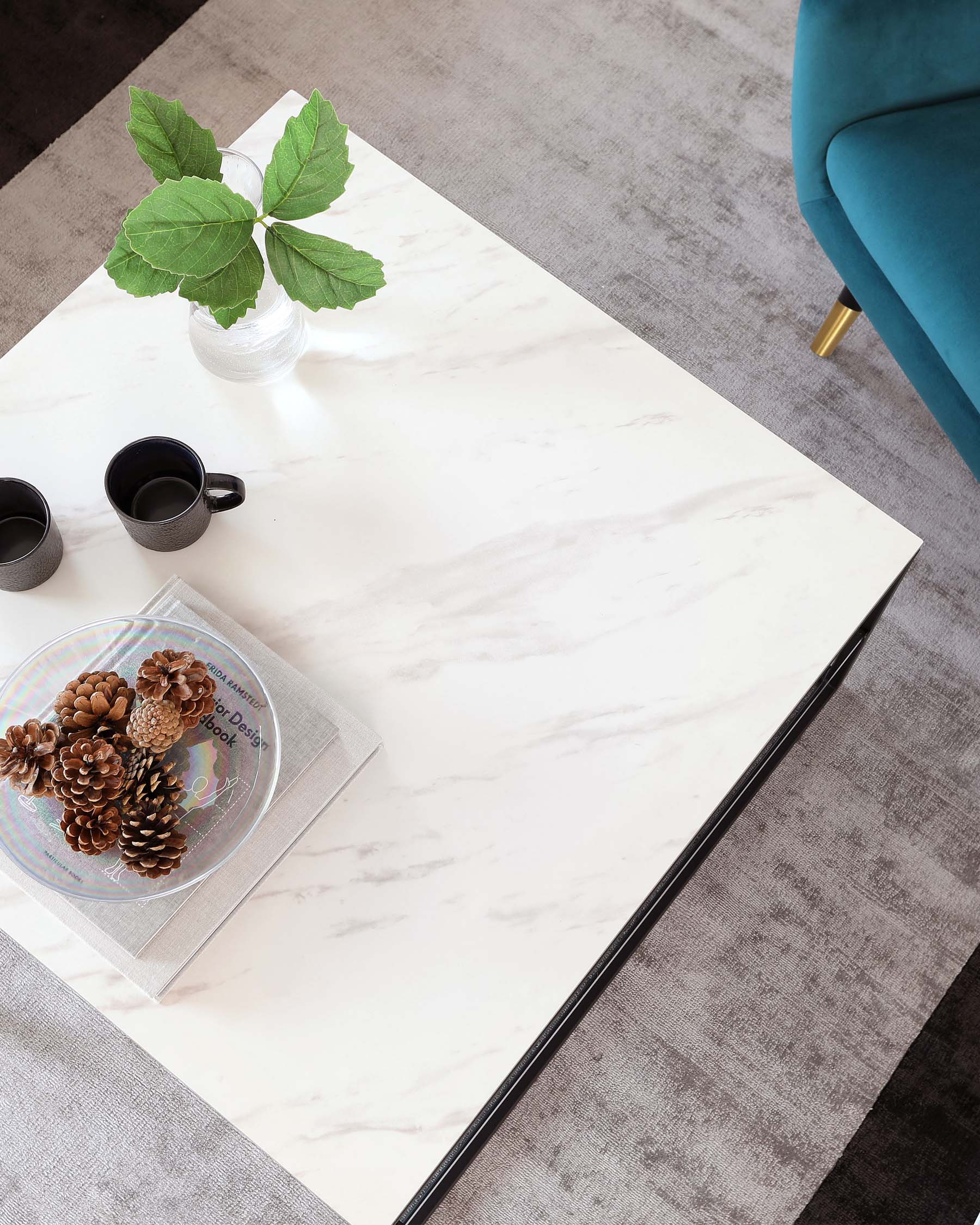 Modern marble-patterned coffee table with a sleek white surface and black metal edges, accompanied by a portion of a teal upholstered armchair with a gold-toned leg peeking from the edge, all set upon a grey textured area rug.