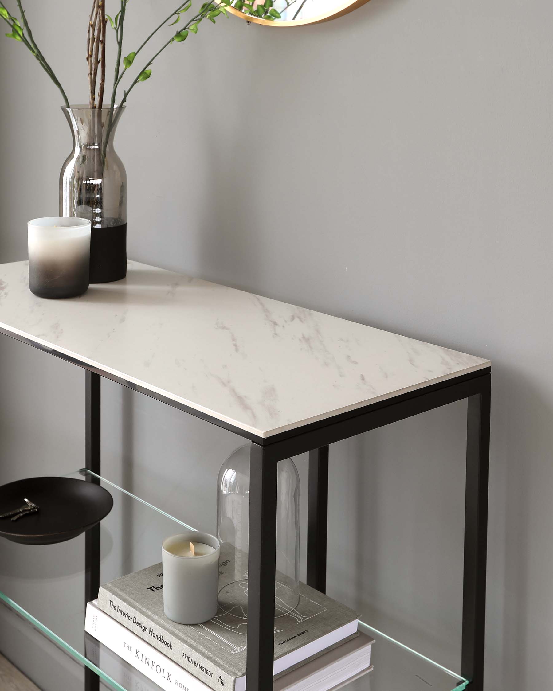 Modern console table with a faux marble top and black metal frame, featuring two open shelves with a glass bottom on the lower shelf and decorative items such as books, a vase with greenery, and candles.