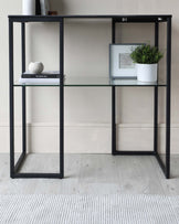 Modern minimalist shelf with a black metal frame and clear glass shelves, featuring two sections with a total of four levels for storage or display. The shelf is styled with a few decorative items such as books, a framed picture, and a plant in a white textured pot. The furniture stands on a light wooden floor with a white and grey area rug beneath it, set against a cream-colored wall.