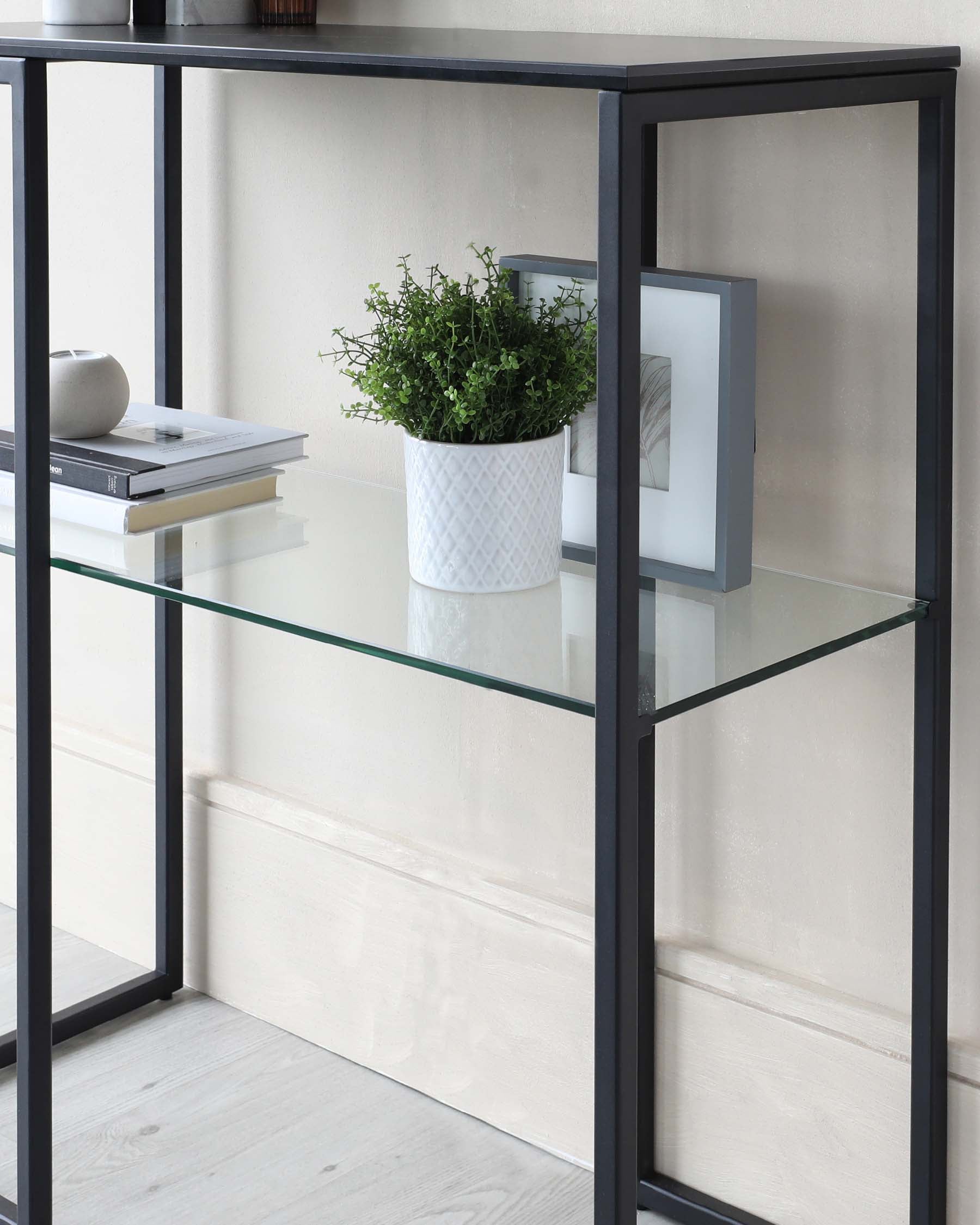 Modern metal-framed console table with two-tiered clear glass shelves, displaying decorative items and a framed mirror leaning against the wall.