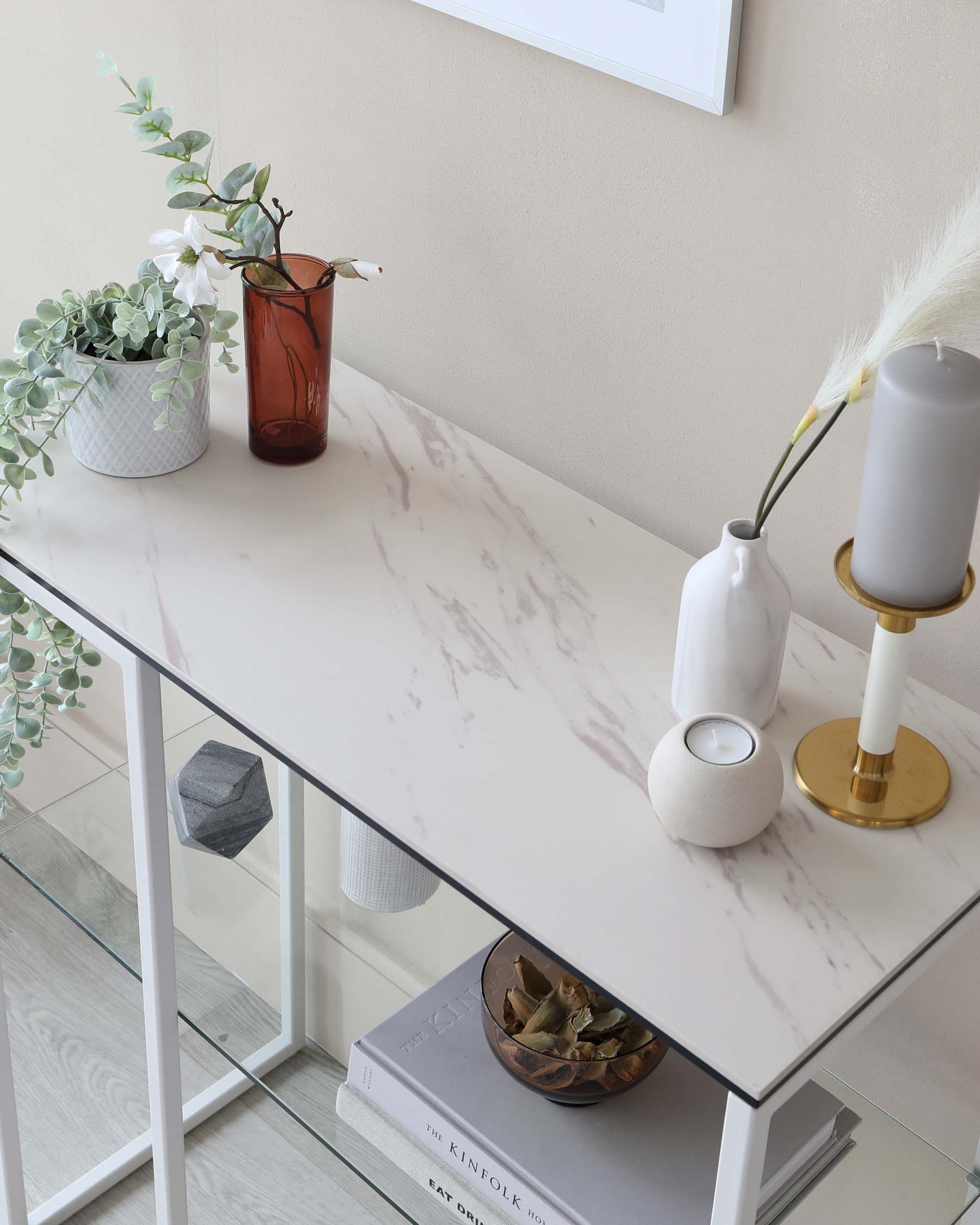 Elegant modern console table with a faux marble tabletop and a minimalist white metal frame. The table is accessorized with decorative items, including a white vase with pampas grass, a candle on a brass stand, a small textured vase, and plant pots. Books are neatly stacked underneath, adding to the sophisticated styling of the space.