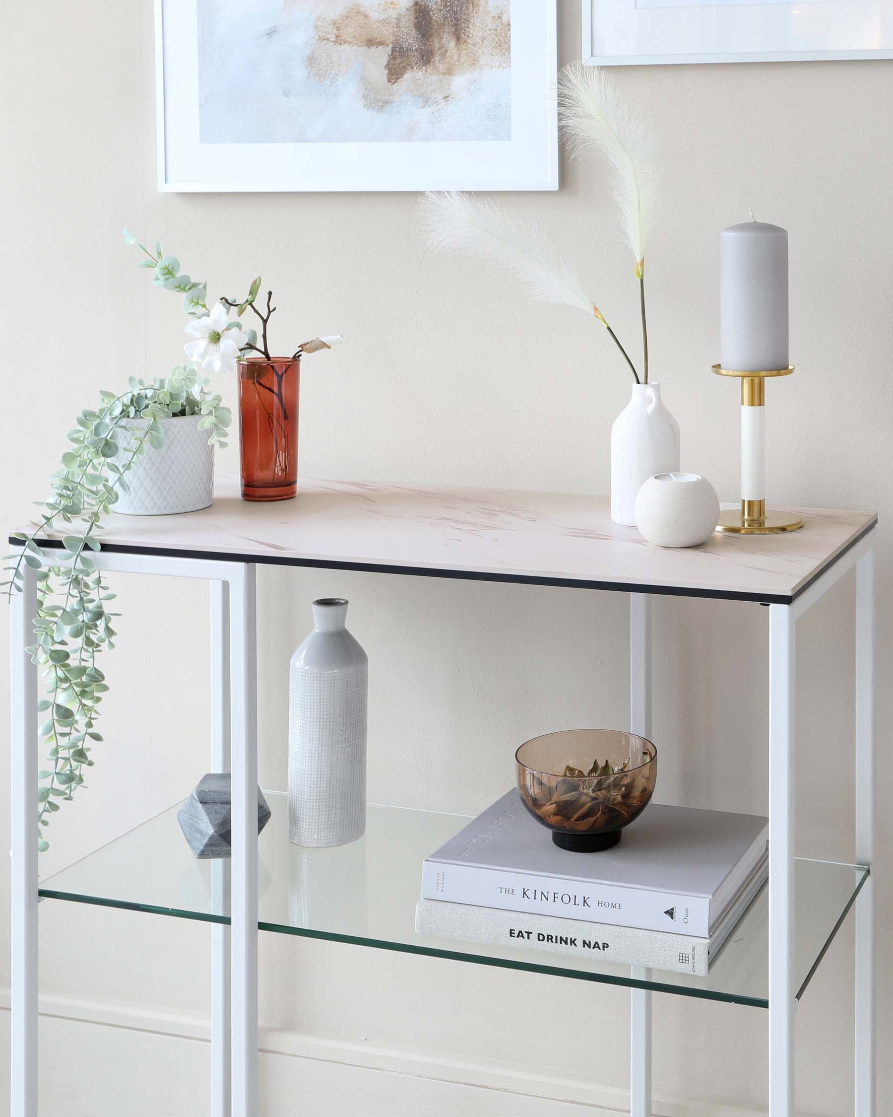 A sleek, contemporary console table with a white marble top and a minimalist metal frame structure finished in matte gold. Underneath, a clear glass shelf offers extra display space.