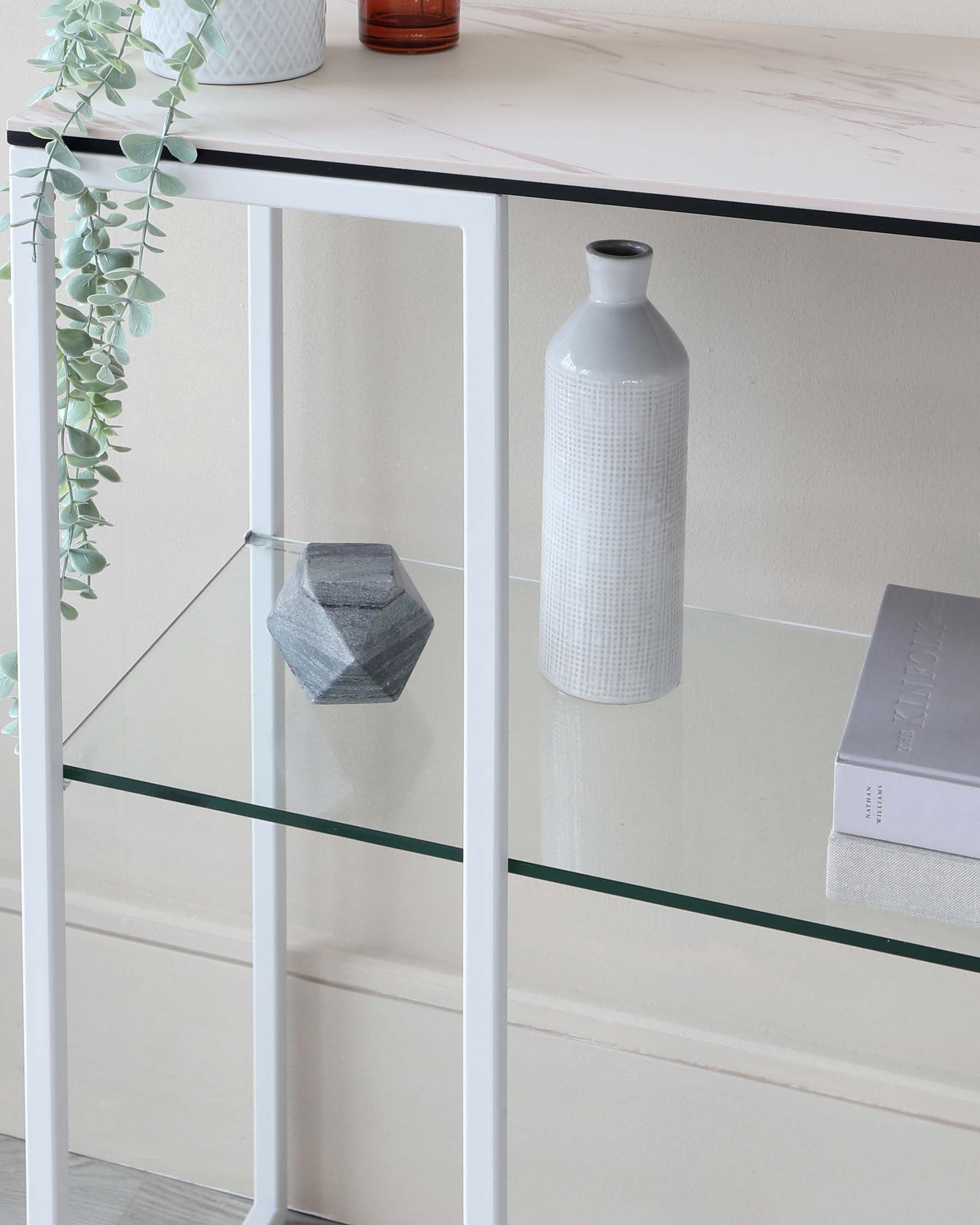 Elegant console table featuring a white frame with thin, straight legs and a marble-patterned tabletop. Below the main top is a secondary glass shelf, displaying a minimalistic grey decorative stone and a book. A tall white textured vase and a potted green plant with trailing foliage accentuate the tabletop, complementing the modern and airy aesthetic.