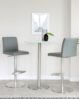 Modern furniture set featuring two grey adjustable-height bar stools with footrests and a round glass-top bistro table on a single sleek metal pedestal base.