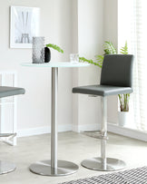 A contemporary round glass-top bistro table with a metallic pedestal base, accompanied by a modern adjustable bar stool featuring a grey seat and backrest, with a matching metallic base.