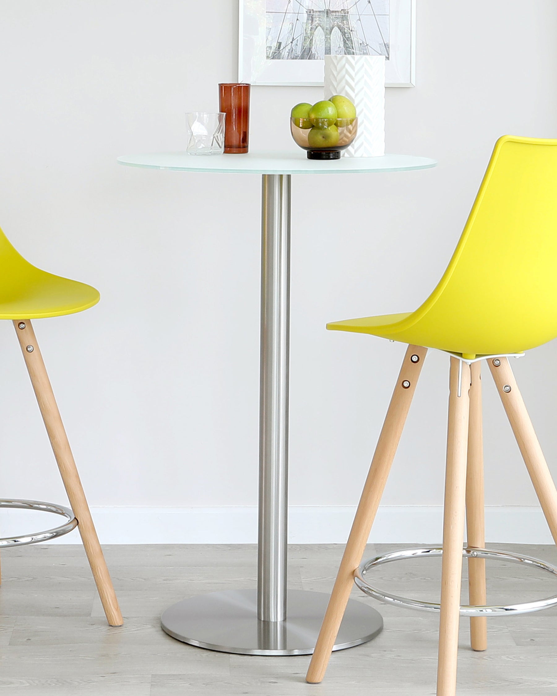 Contemporary round bistro table with a frosted glass top and a sleek, silver metal pedestal base complemented by two modern chairs with yellow moulded plastic seats and wooden legs with metal frame supports.