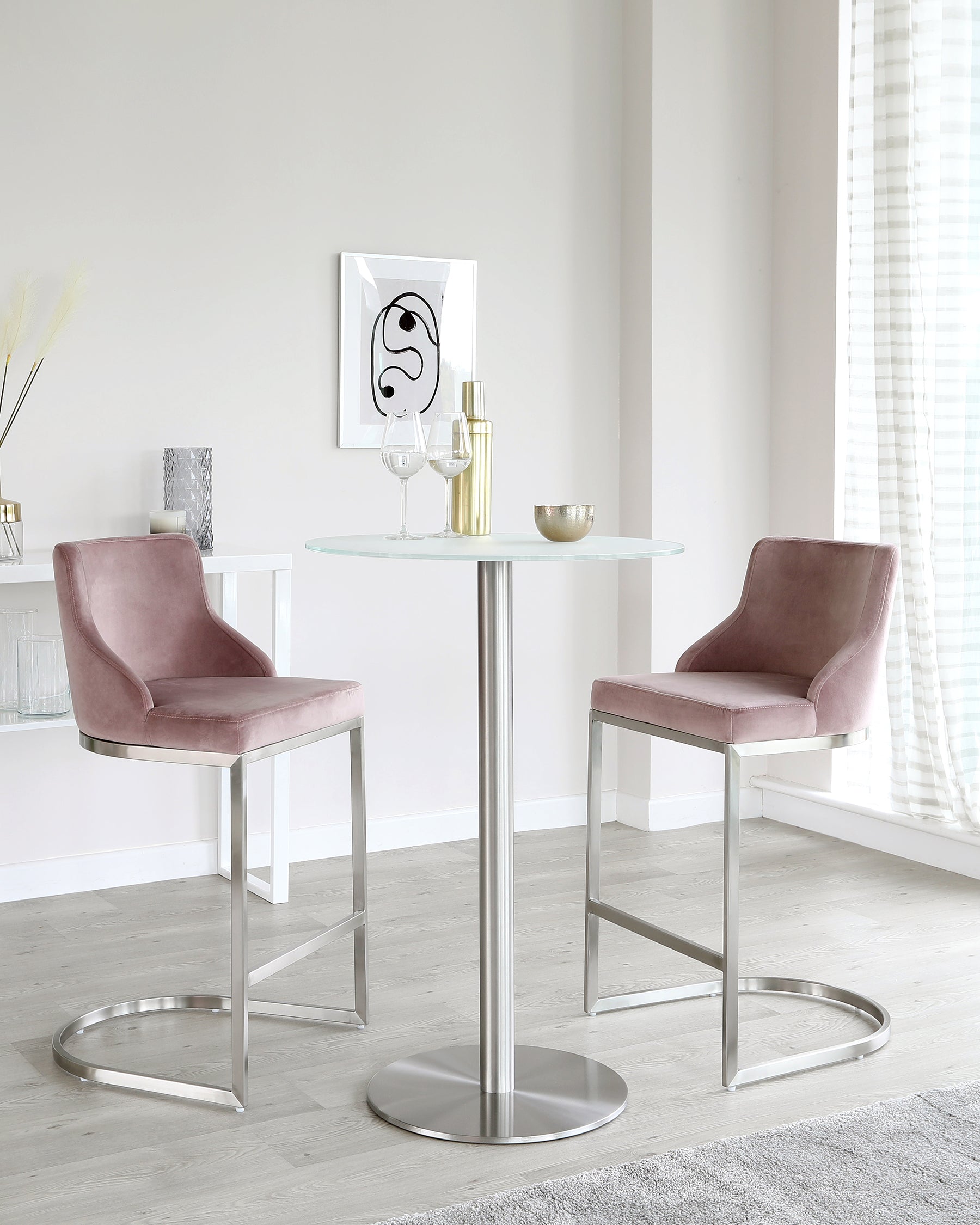 A modern dining set with two mauve velvet upholstered bar chairs featuring a sleek metal frame with a footrest, and a round glass-top table standing on a central polished metal pedestal.