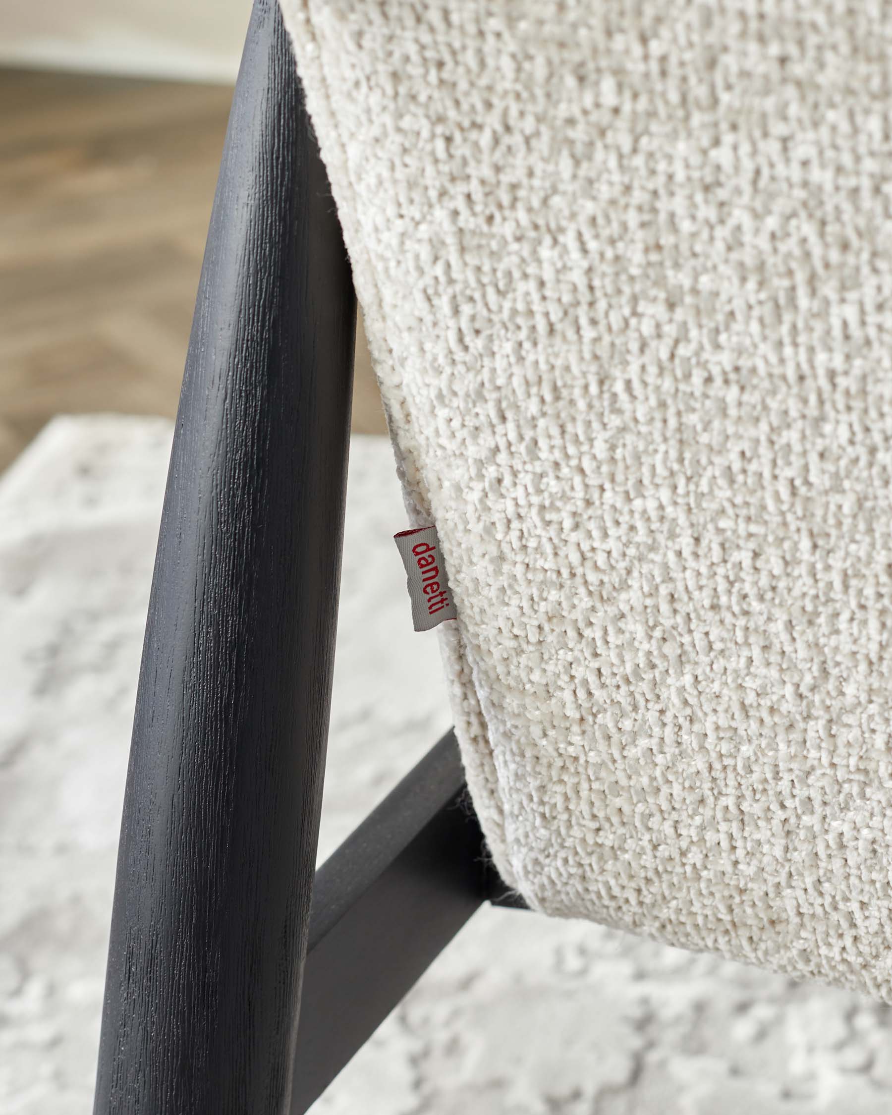 A close-up of a modern chair with a black wooden frame and textured off-white upholstery fabric. A small red tag with branding is visible on the side of the fabric. The chair is partially seen, and there's a focus on the material and the wooden frame's textures, with a blurred background that hints at an indoor setting.