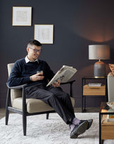 Modern minimalist armchair with a cream upholstery and dark wooden frame, paired with a sleek side table featuring dark metal legs and a wooden drawer, complemented by a contemporary table lamp with a cylindrical ribbed base and a grey drum shade.