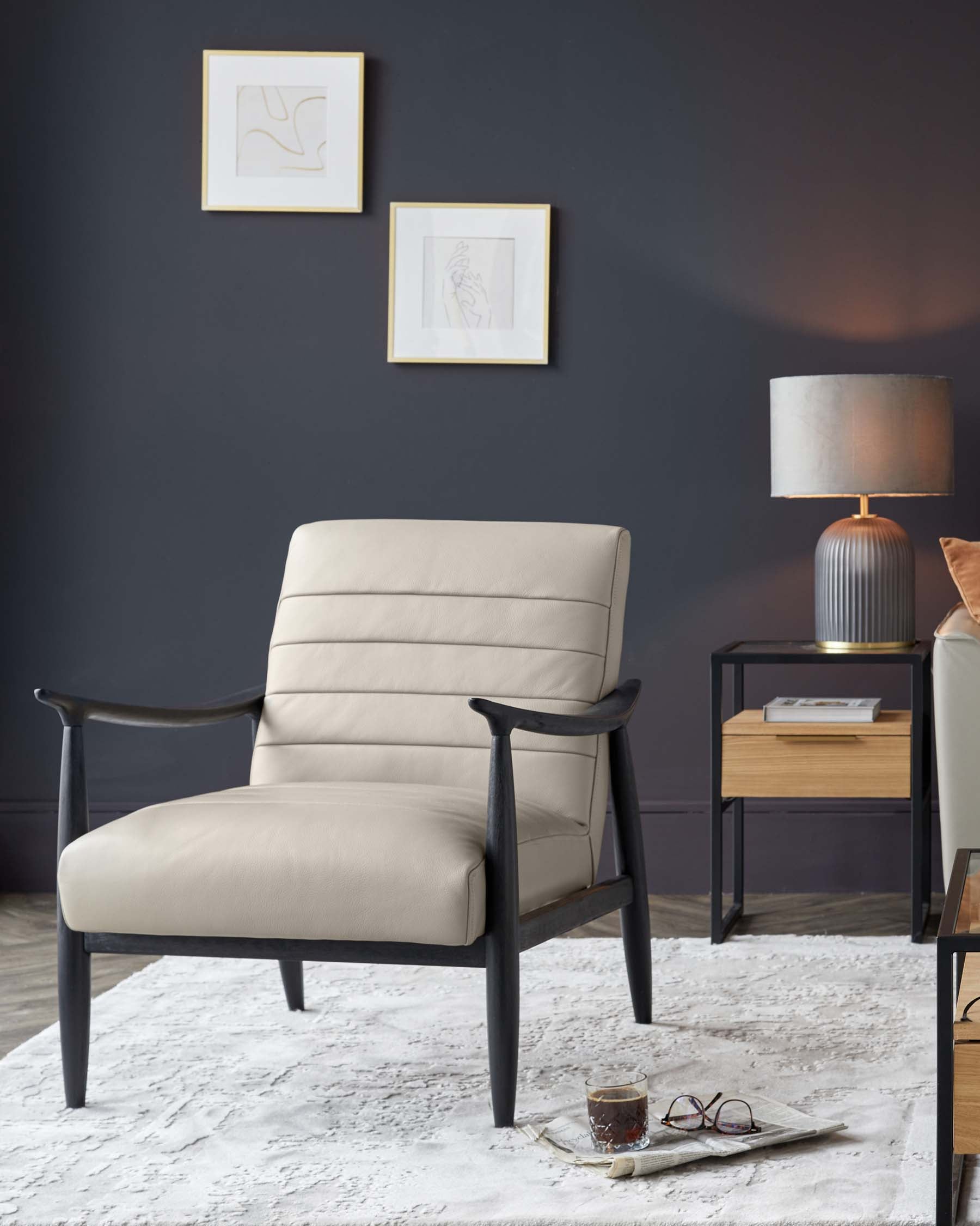 Modern armchair with light beige upholstery and dark wooden frame, beside a contemporary style side table with a natural wood finish drawer and black metal legs, atop a textured white area rug. A table lamp with a ribbed base and grey shade adorns the side table.