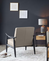 A contemporary taupe armchair with black wooden frame and angled armrests, accompanied by a small wooden side table with an open shelf and a rectangular tabletop, complemented by a textured grey lamp with a cylindrical shade, all staged on a soft white area rug against a dark grey wall with two framed abstract artworks.