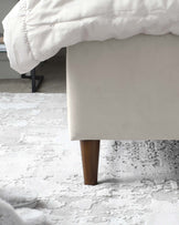 Contemporary upholstered bed frame corner with visible tapered wooden leg on a textured white area rug.