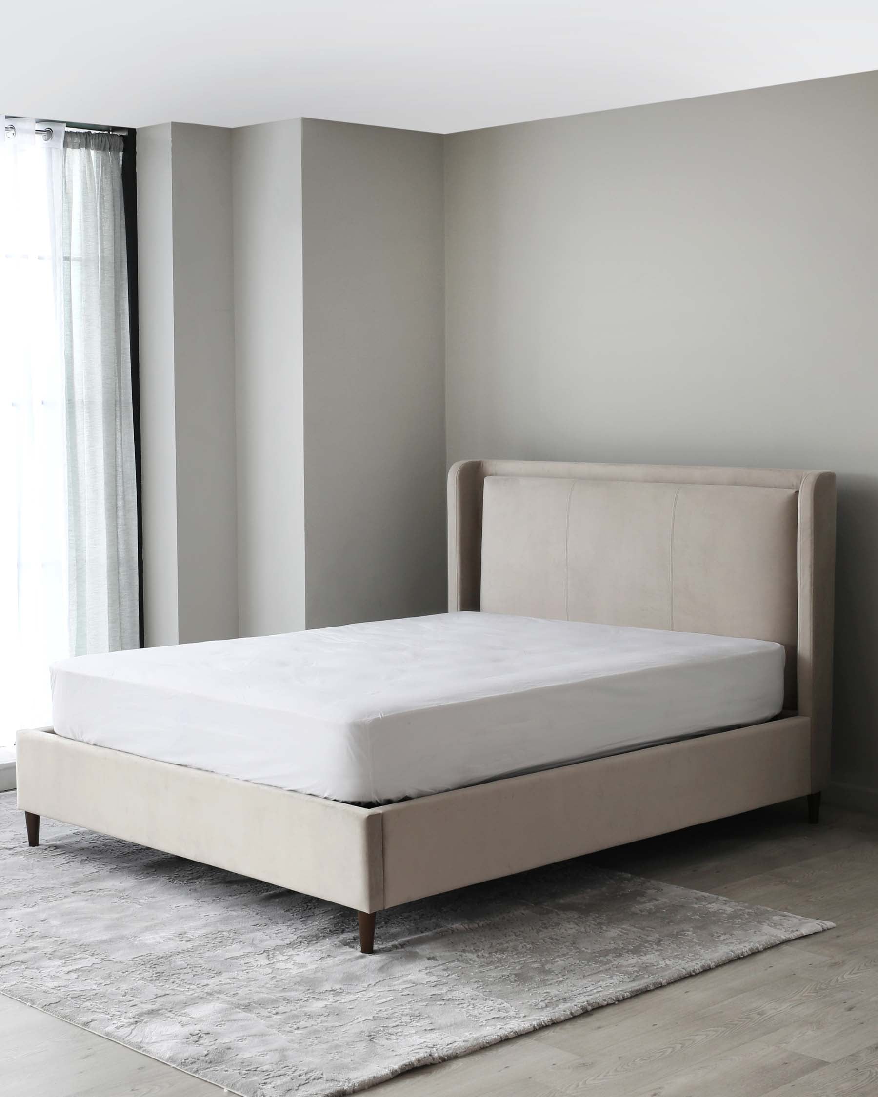 Elegant modern bed with a simple headboard and upholstered frame in a light beige fabric, set upon dark wooden legs, accompanied by a white fitted sheet on the mattress, all positioned on a textured grey area rug.