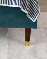 Elegant dark green upholstered ottoman with a plush finish and tapered dark wooden legs accented with gold-coloured tips, partially draped with a striped blue and white blanket, on a light-coloured carpeted floor.