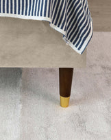 Beige upholstered sofa with dark wooden legs capped with a gold tip, partially draped with a blue and white striped throw blanket, on a light grey textured carpet.