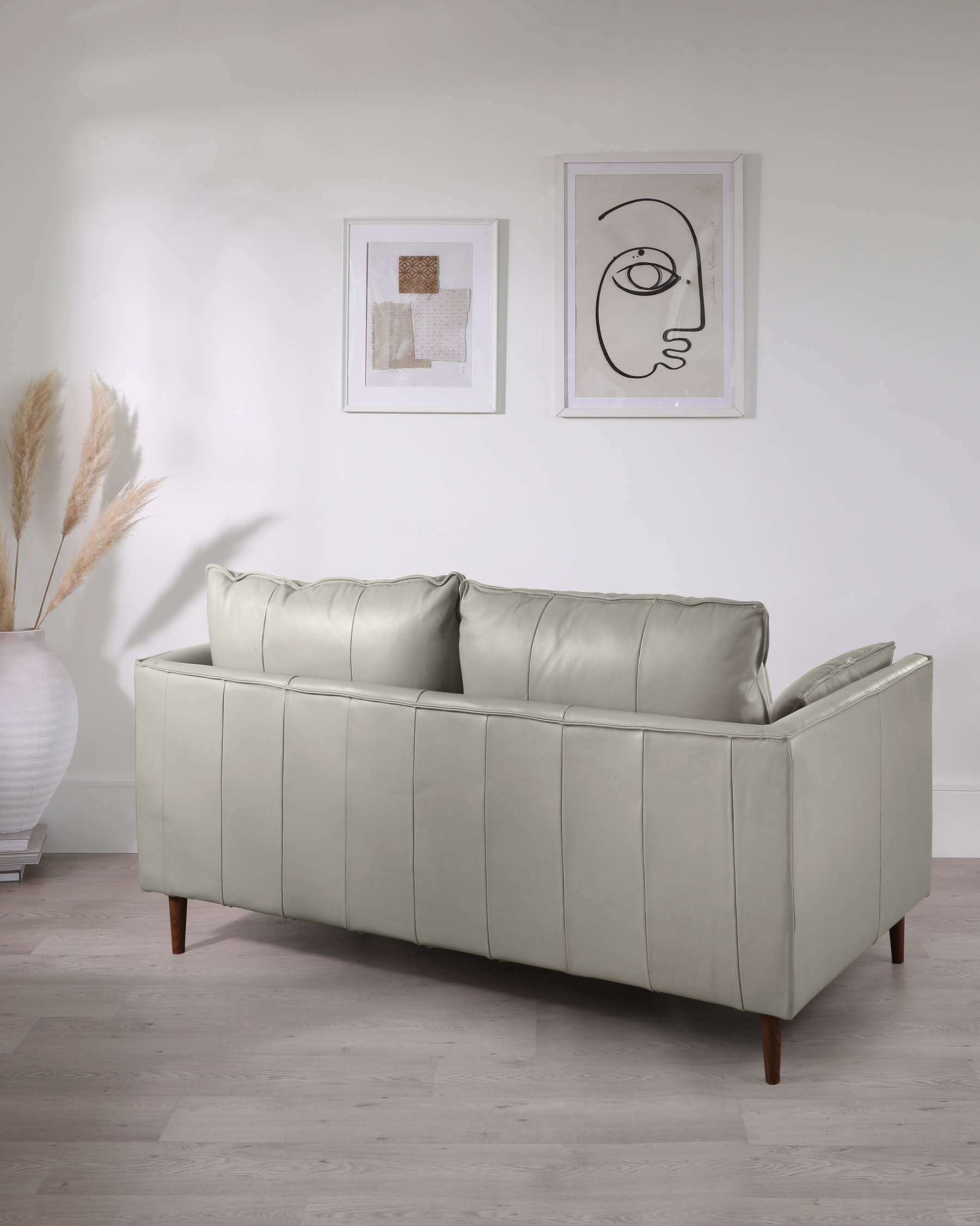 Elegant light grey leather three-seater sofa with clean lines and wooden tapered legs on a light hardwood floor, set against a neutral wall with minimalist framed artwork, accompanied by a vase of pampas grass on a small end table.