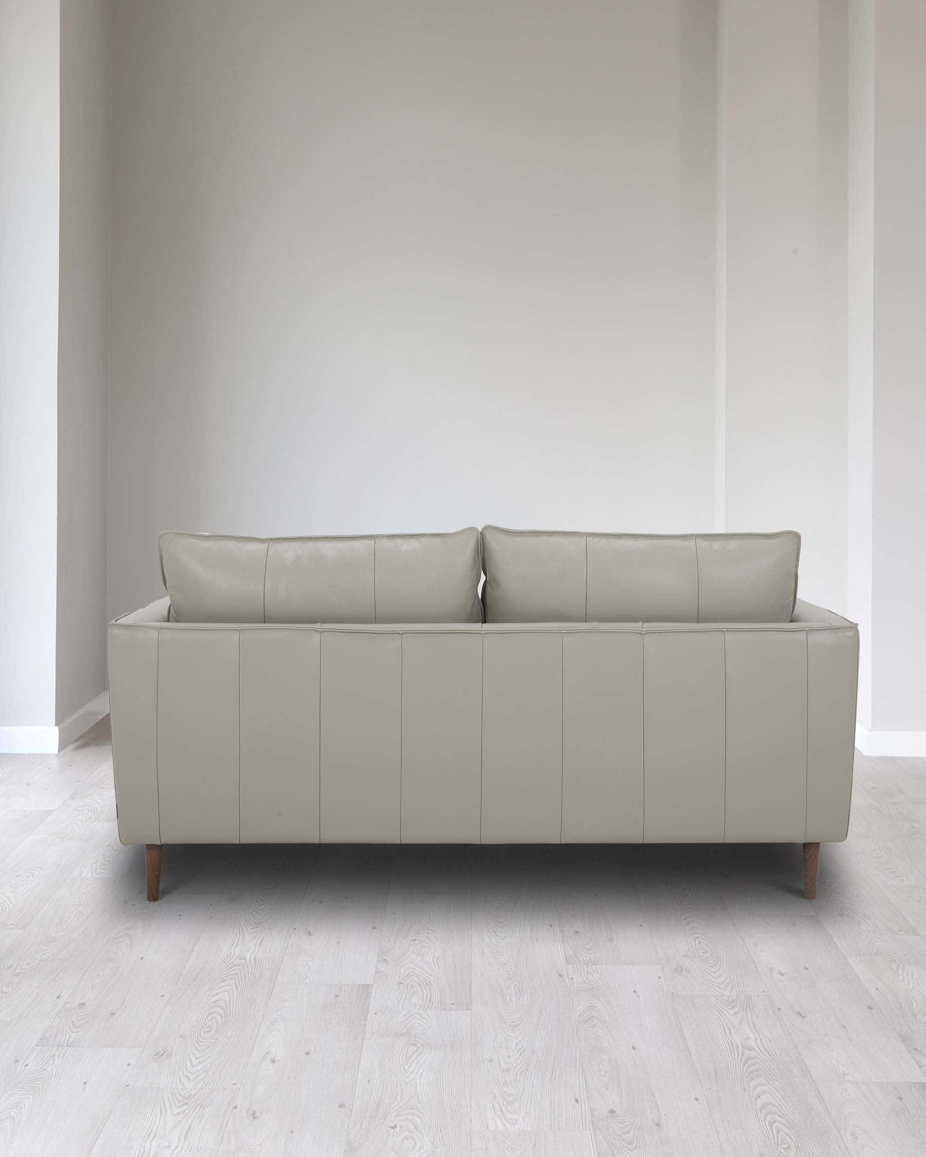 Modern light grey three-seater sofa with a minimalist design, featuring clean lines, square arms, and two plush back cushions, set on angled wooden legs against a neutral wall and light wooden flooring.