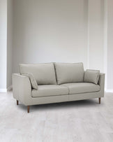 A modern three-seater sofa with a minimalist design, featuring straight lines and smooth light grey upholstery. The couch stands on four wooden tapered legs and includes three large back cushions and one smaller side cushion for added comfort. It is positioned on a light wooden floor against a neutral white wall which emphasizes its sleek silhouette.