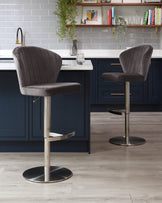 Two modern bar stools with elegant grey velvet upholstery and vertical channel tufting. Each stool features a curved back and seat design, mounted on a sleek, adjustable-height metal pedestal base with a built-in footrest and circular floor plate. The stools are positioned at a kitchen island with a navy blue base and white countertop, complementing a contemporary interior.