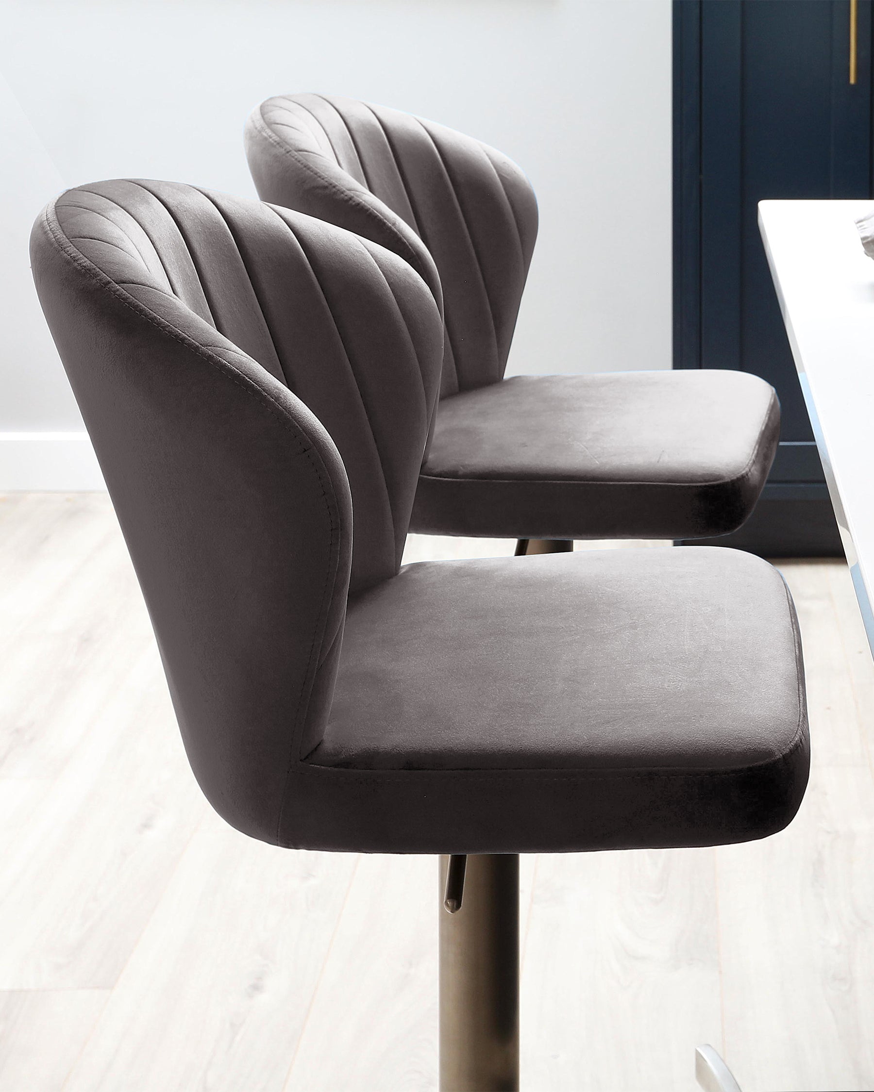 Two contemporary dark grey upholstered dining chairs with vertical channel tufting on the high backs and smooth cushioned seats. Both chairs feature a sleek, armless design and rest on slender metallic legs with a round base, suggesting a swivel functionality. The chairs are set on a light wooden floor against a minimalist backdrop with a subtle contrast of a dark-coloured door.