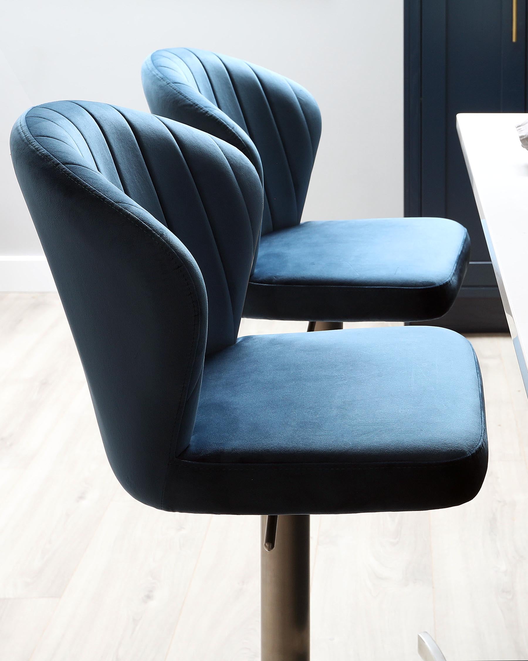 Two modern bar stools with deep blue velvet upholstery and channel tufting on the backrest, featuring curved wingback design and black metal base with footrest.