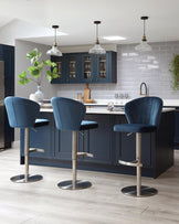 Three modern bar stools with plush velvet upholstery in a rich blue, featuring curved backrests and cushioned seats. The stools have sleek metallic bases with built-in footrests and gas-lift height adjustment mechanisms.