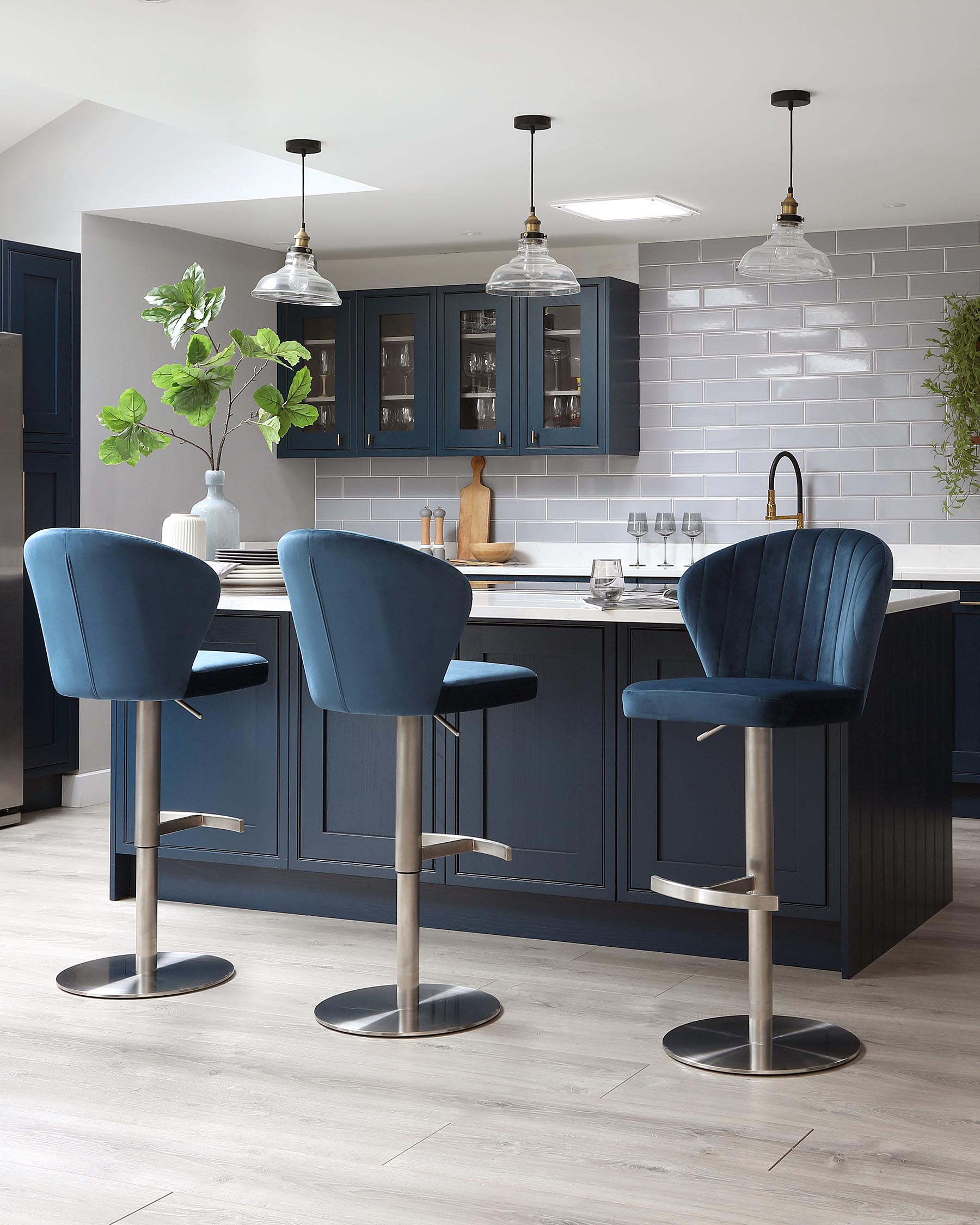 Three modern bar stools with plush velvet upholstery in a rich blue, featuring curved backrests and cushioned seats. The stools have sleek metallic bases with built-in footrests and gas-lift height adjustment mechanisms.