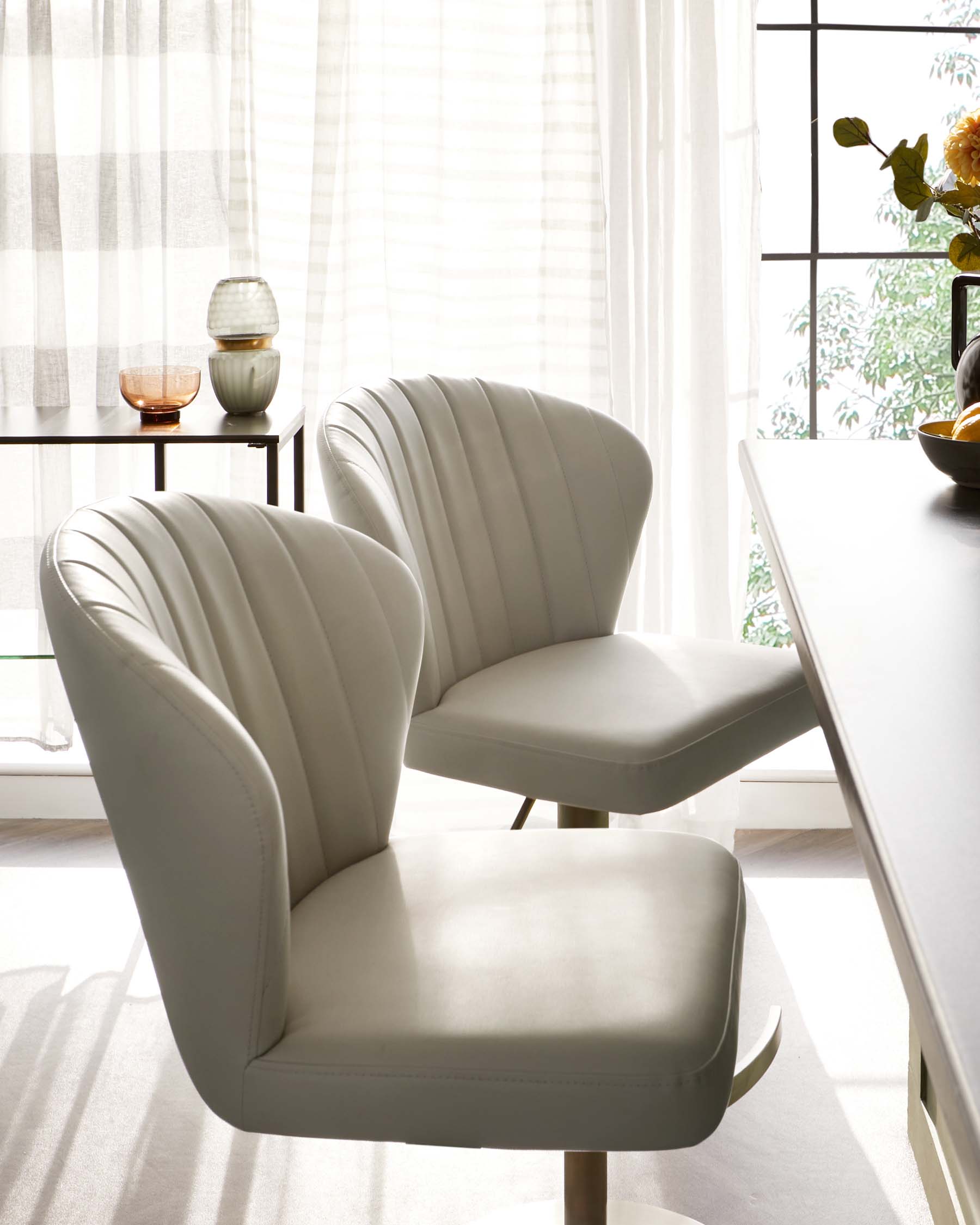 Two modern light grey upholstered dining chairs with vertical channel tufting and splayed wooden legs, set in a bright room with sheer curtains and natural light. A small round table with decorative items is partially visible in the background.