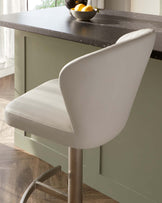 Elegant modern bar stool with a curvaceous backrest and seat upholstered in off-white faux leather, featuring a sleek brushed metal pedestal with footrest and contrast stitching detail, positioned at a kitchen island with a dark stone countertop.