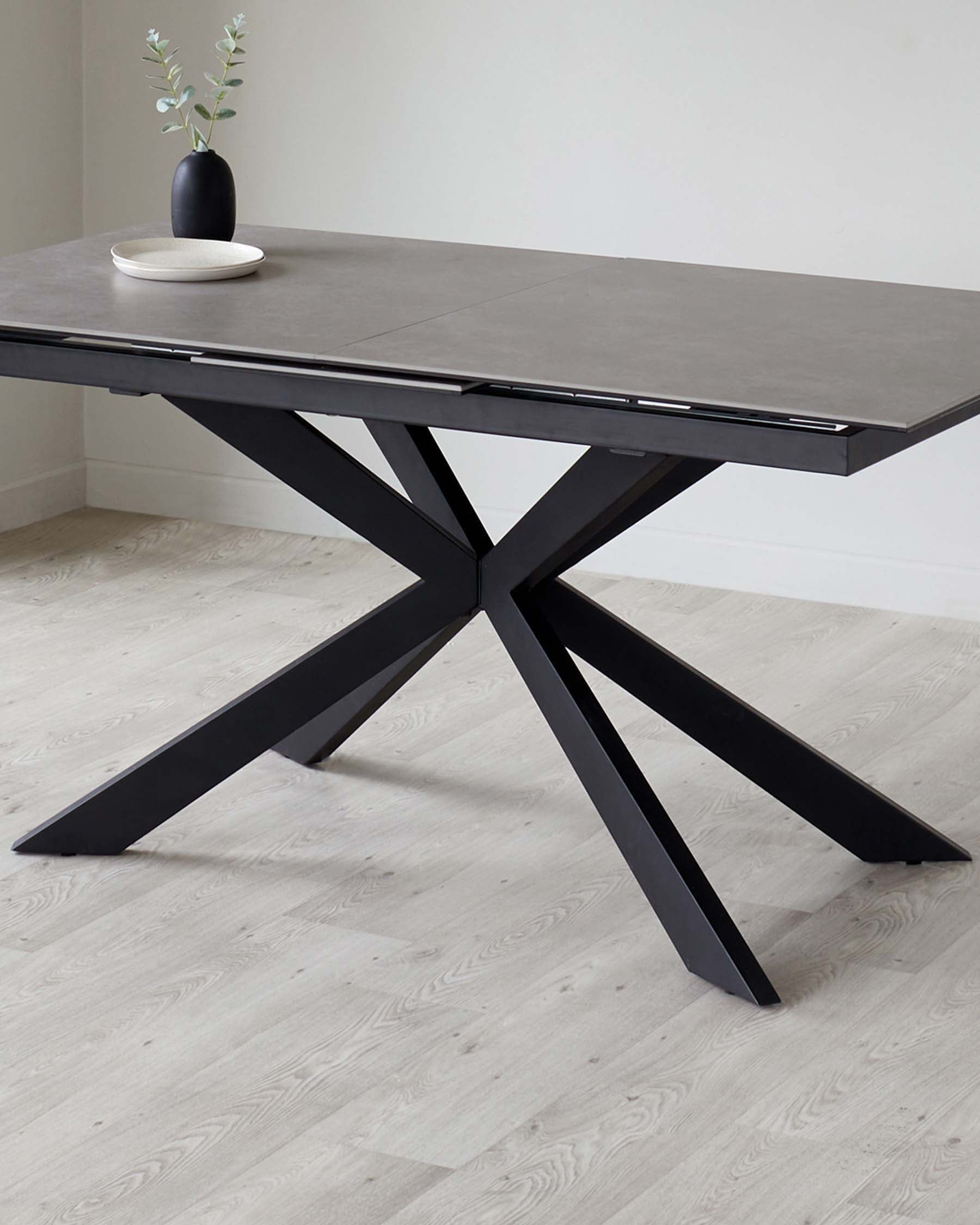 Contemporary dark grey extendable dining table with a distinctive X-base design in a black finish, showcasing a smooth rectangular top with clean lines, perfect for modern interiors. A simple vase with greenery is displayed on the table as decor.