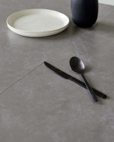 No furniture is visible in this image, only a cream-colored ceramic plate, a dark vase, and a black spoon and knife on a grey stone surface.