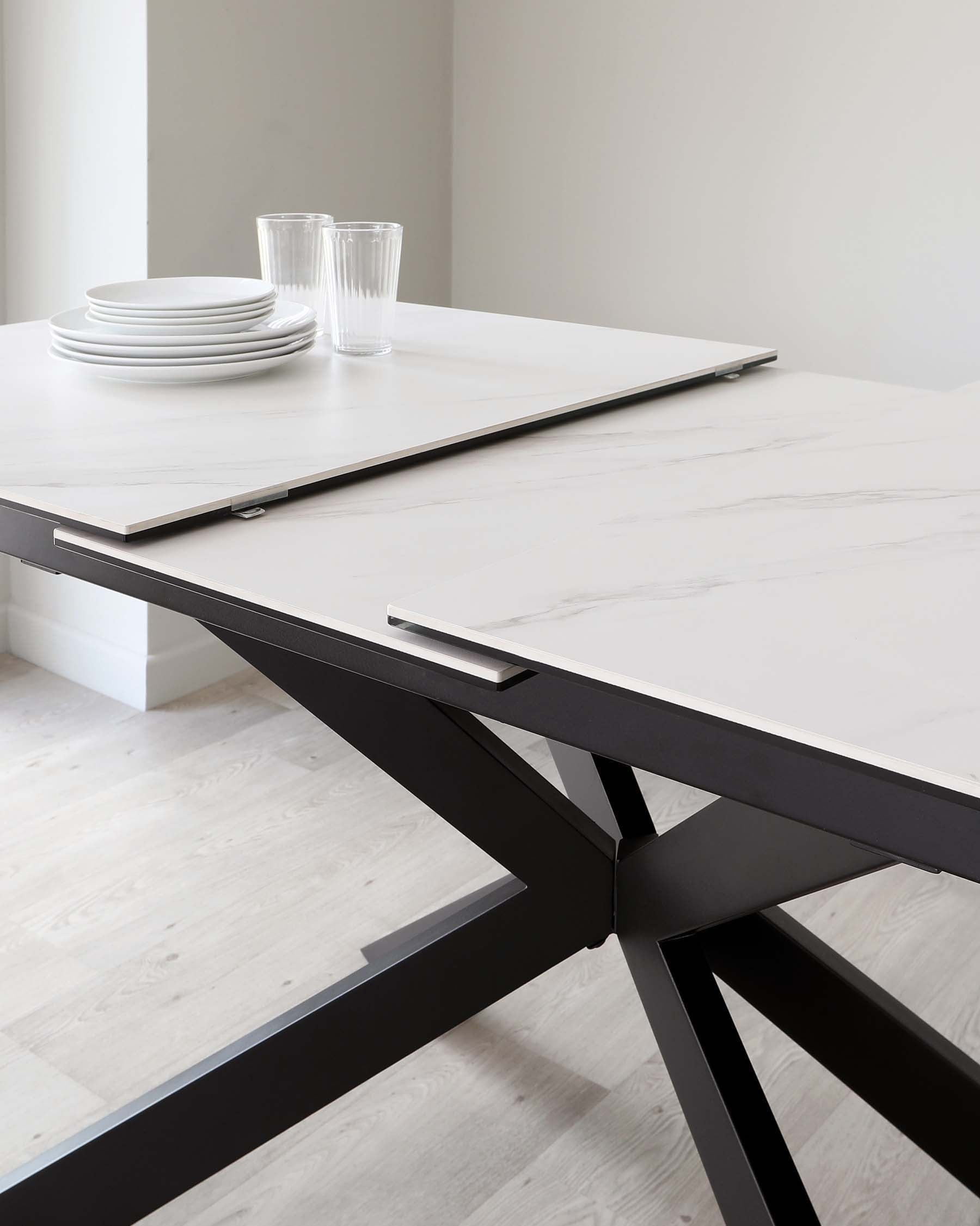 A modern dining table with a white marble tabletop and a bold, black geometric base. The table is partially set with white ceramic plates and clear glassware.