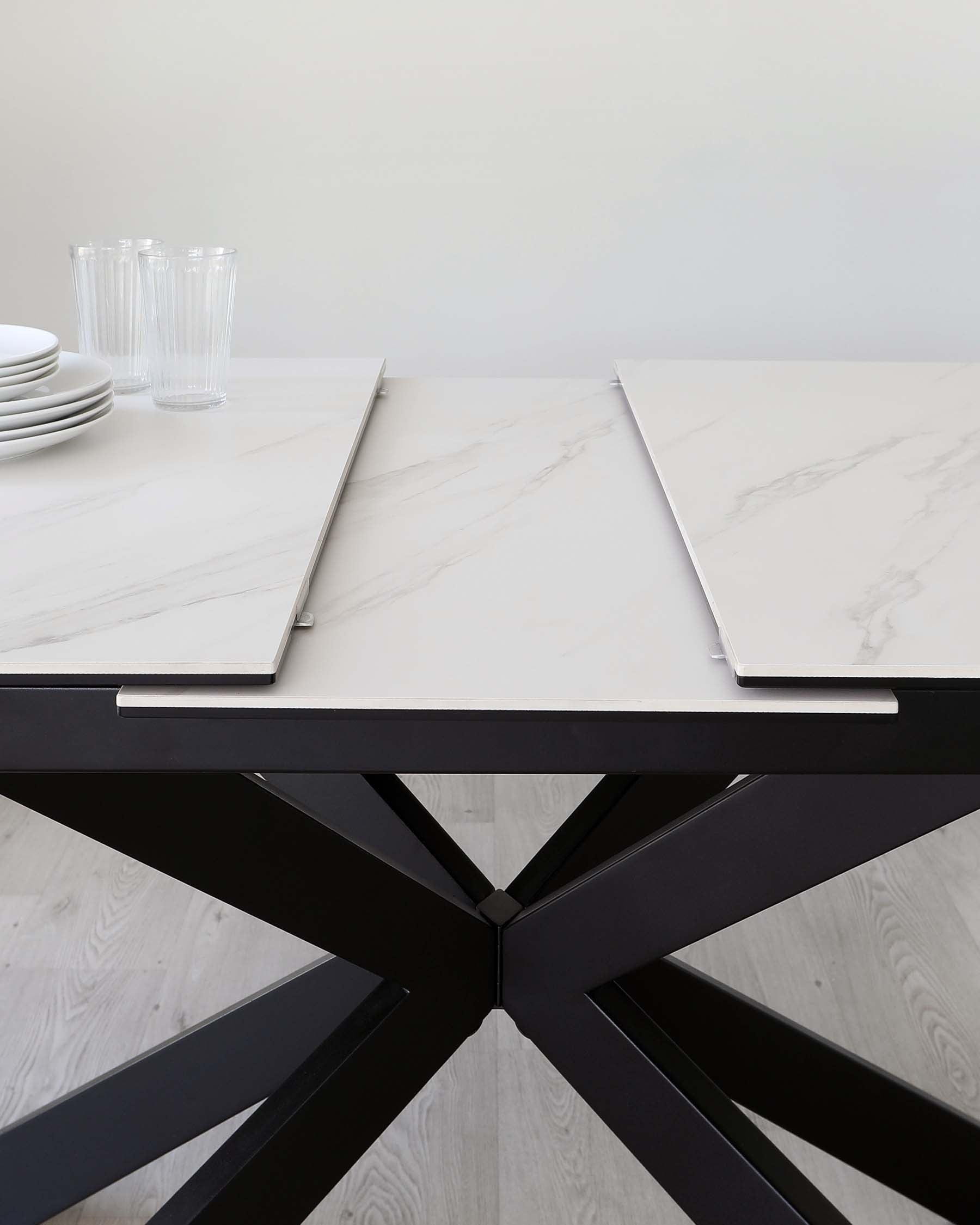 Modern dining table with a white marble top supported by a striking black starburst-style metal base. The tabletop has sleek straight edges, and on top of the table are stacks of white plates and clear glass tumblers, suggesting a minimalist and elegant setting.