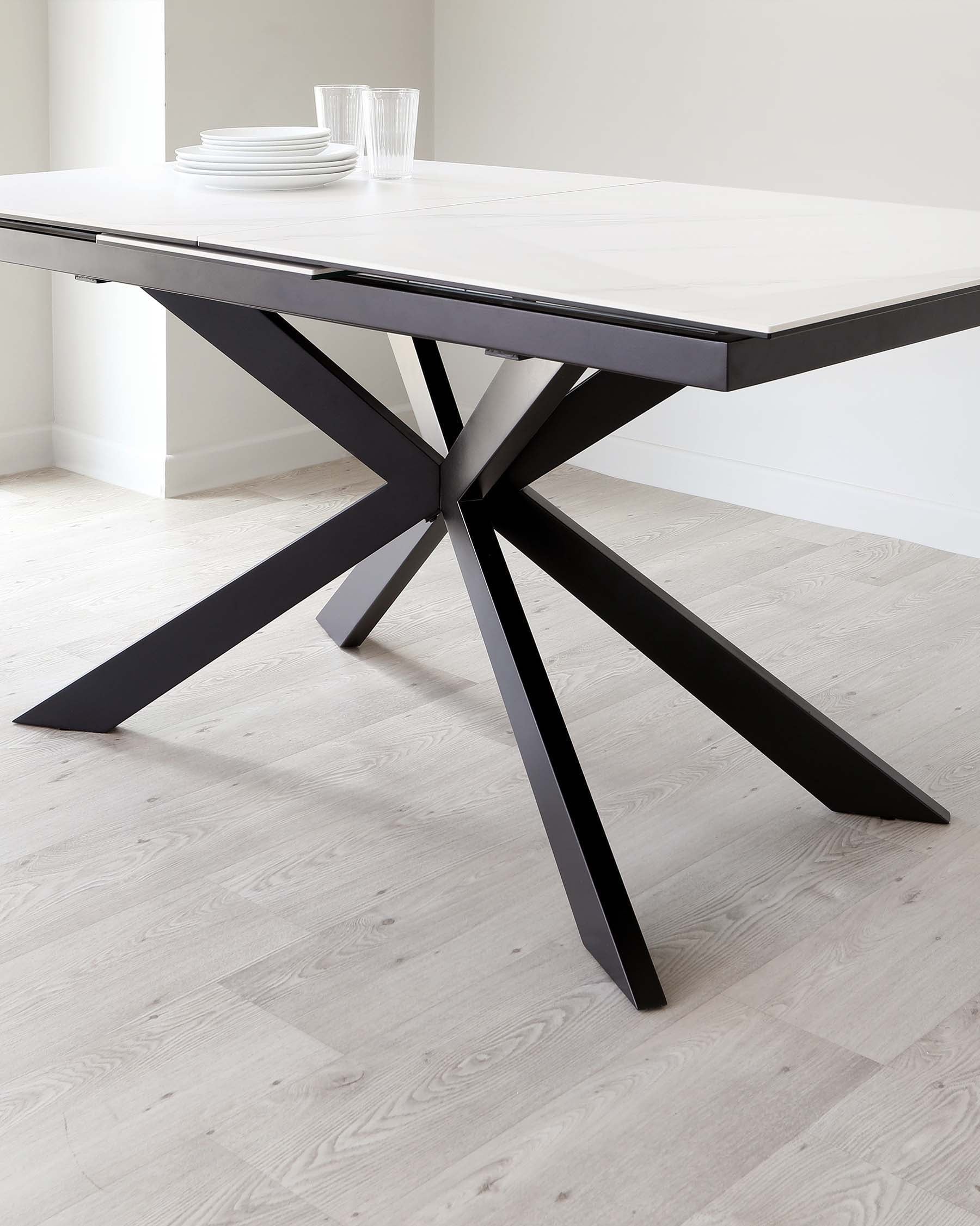 Modern extendable dining table with a matte white top and a black star-shaped base on a light hardwood floor. The table is partially extended and adorned with a simple white dish set and clear glasses.