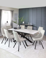 A modern dining room set featuring a large, rectangular, white marble table with a geometric black base. Surrounding the table are six mid-century-inspired chairs with curved, white upholstered seats and black tapered legs. A simple black console table sits against a dark panelled wall in the background.