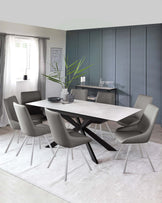 Modern dining room set with a white rectangular table featuring a sleek tabletop and contrasting black angular legs. Accompanied by six minimalist grey upholstered chairs with slender metal legs. The setup is on a soft white area rug and accessorized with a grey vase holding greenery, black dishes, and clear glassware on a slim black console table in the background.