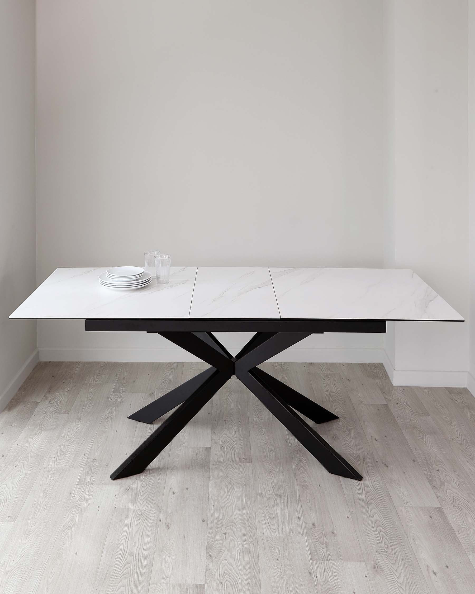 A modern rectangular dining table with a white marble-like tabletop and a striking black crisscrossed metal base, placed on a light wooden floor. The table is set with a stack of white plates and several clear glasses. The room has white walls and is well-lit, providing a contemporary and minimalist aesthetic.