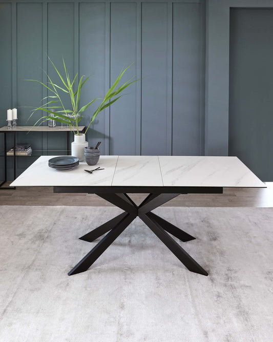 Modern rectangular dining table with a white marble tabletop and a bold black X-shaped metal base, accompanied by a minimalist metal shelf unit with decorative items against a muted teal wall.