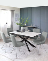 Modern dining room featuring a white rectangular table with a black crisscross leg design and six sleek, grey upholstered chairs with metal legs, set on a light grey area rug.