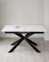 Modern rectangular dining table with a white marble tabletop and a bold black X-shaped base, set on a light hardwood floor with simple white dinnerware on top.