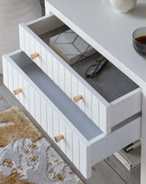 Modern white console table with clean lines, featuring two stacked drawers with vertical groove detailing. Drawers are accented with minimalist wooden knobs, contrasting the crisp white finish. A low, open shelf runs the length of the table just below the top surface, providing additional display or storage space. The piece is staged on a grey and cream area rug, suggesting a contemporary home setting.