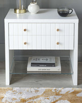 Modern white nightstand with three drawers, featuring horizontal line detailing and round wooden knobs. The nightstand has a lower shelf with a glass surface, ideal for books or decor items, and stands on square metal legs. A clear glass coffee cup and a round white vase with a gold accent are placed on top of the nightstand.