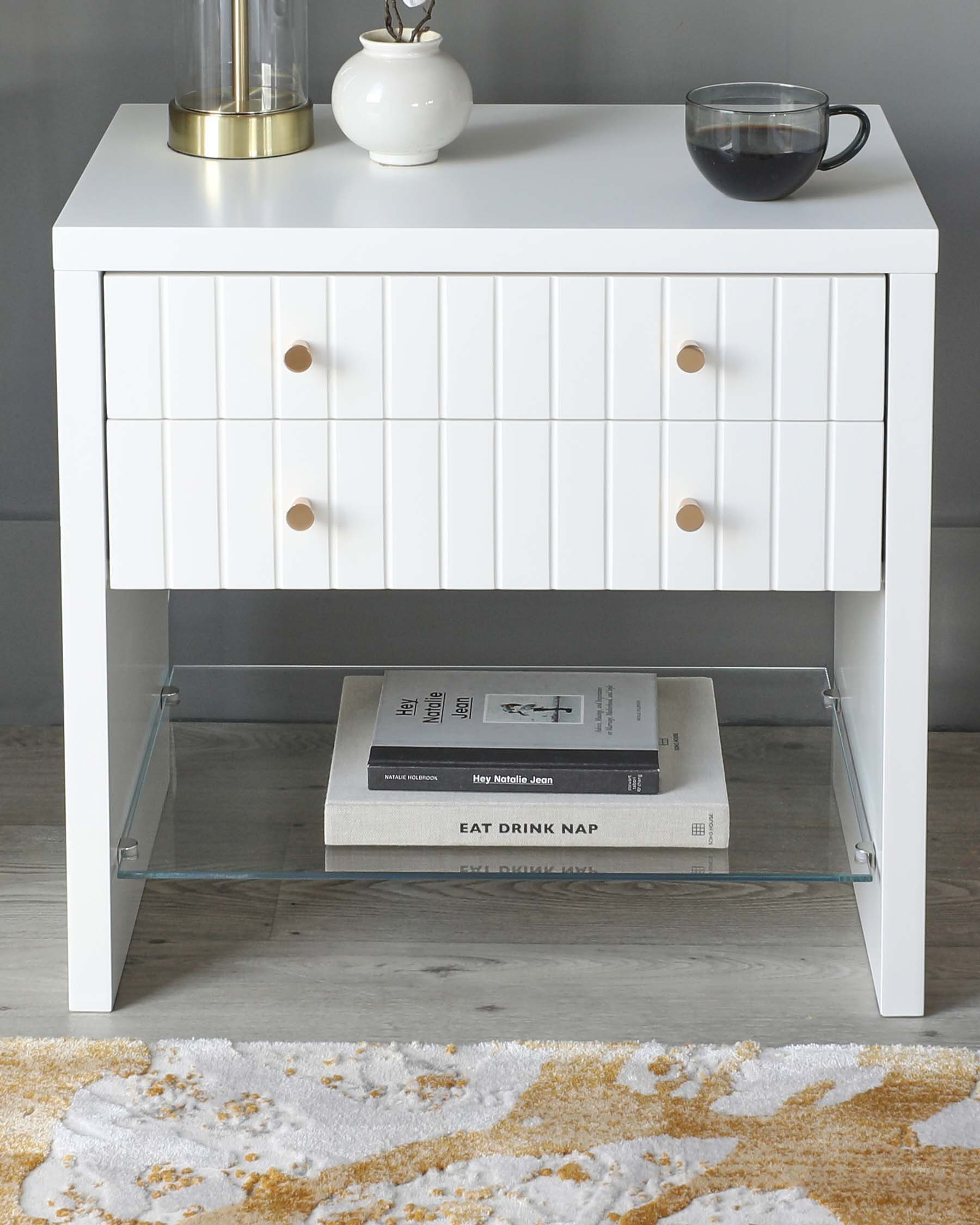 Modern white nightstand with three drawers, featuring horizontal line detailing and round wooden knobs. The nightstand has a lower shelf with a glass surface, ideal for books or decor items, and stands on square metal legs. A clear glass coffee cup and a round white vase with a gold accent are placed on top of the nightstand.