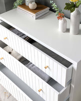 Modern white dresser with clean lines and a minimalist design, featuring multiple drawers with horizontal grooves and wooden knobs. The dresser top is styled with decorative items including books, a spherical vase, and ceramic vases with flowers. The wall behind it is painted a subtle grey, adding to the contemporary aesthetic.