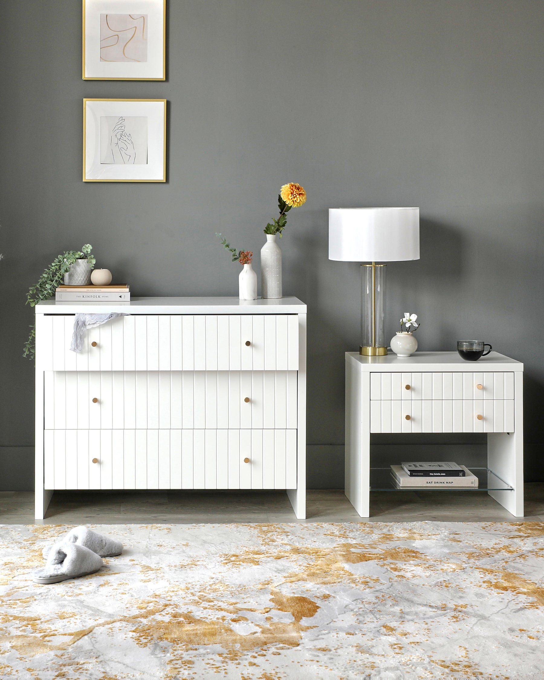 Modern white wooden dresser with vertical beaded drawer fronts and brass knobs, accompanied by a matching side table featuring a drawer and an open shelf beneath. Both pieces set against a grey wall with decorative items on top, standing on an ornate faded rug.