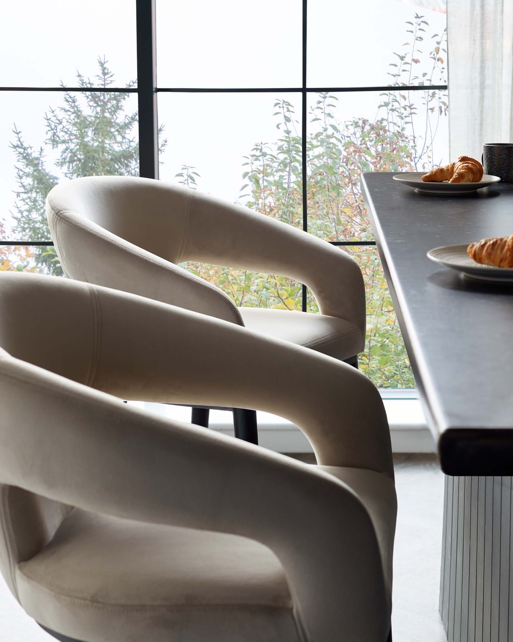 Elegant modern dining chairs with a curved back design, upholstered in a light beige fabric, paired with a contemporary dark table with a fluted texture on its side panel, in a bright room with a large window showcasing a natural view.