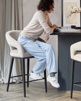 Elegant modern bar stool with a curved, upholstered beige seat and supportive backrest on a sturdy black metal frame with four legs and footrests, paired with a sleek dark countertop bar table.