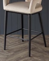 Elegant modern bar stool featuring a sleek black metal frame with four tapered legs and a geometric footrest. The seat and backrest are upholstered in a smooth, light beige fabric, providing a comfortable and stylish seating option. The stool balances contemporary design with classic comfort, perfect for any chic kitchen or home bar setting.